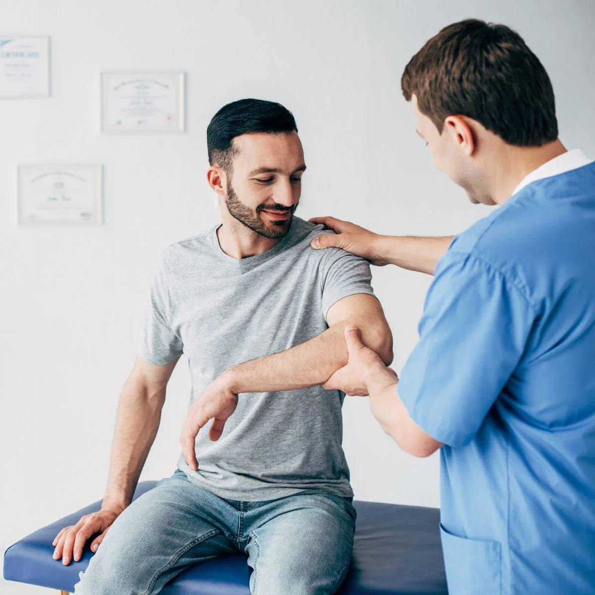 A male physical therapist in blue scrubs is performing a range-of-motion test on a smiling male patient's shoulder and elbow.