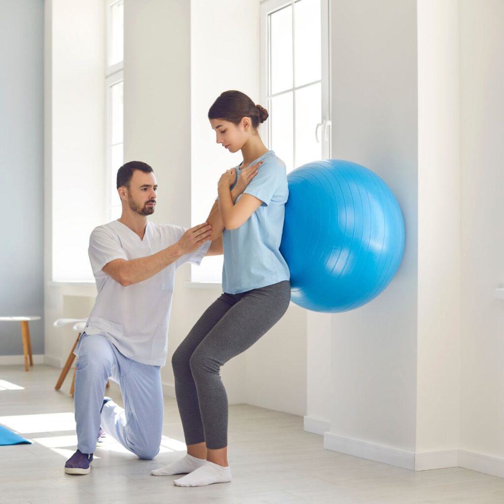 A male physical therapist is guiding a female patient through a wall squat exercise using a large blue stability ball for knee or lower body rehabilitation and strengthening in a bright clinic.